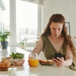 Pregnant woman at breakfast table browsing on smartphone while having a healthy meal indoors.