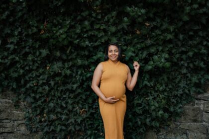 Maternity photo of a woman in autumn, wearing a mustard dress against a leafy backdrop.