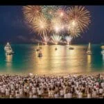 Pessoas vestidas de branco celebrando o Réveillon 2026 na Praia dos Anjos em Arraial do Cabo, com fogos refletindo sobre o mar azul-turquesa e barcos iluminados ao fundo.