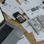 A desk with financial documents, currency, a laptop, and phone calculator.