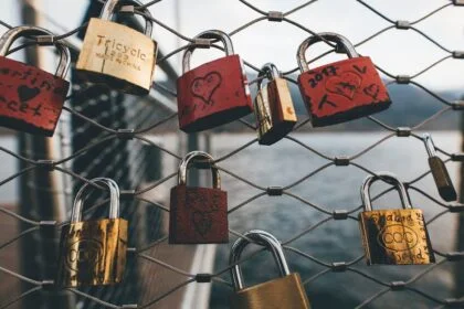 Red and gold padlocks with love messages hang on a chain link fence near water.