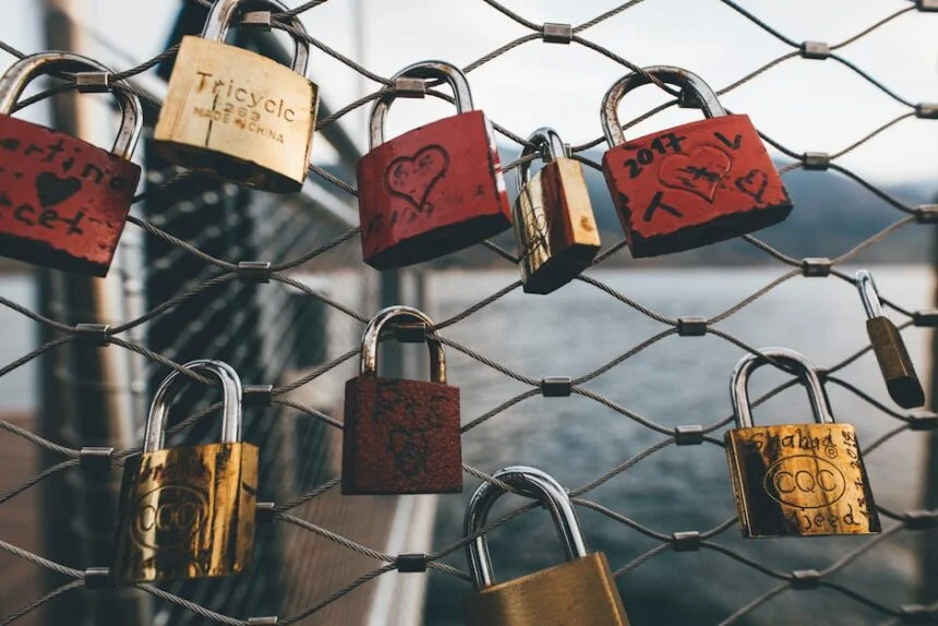 Red and gold padlocks with love messages hang on a chain link fence near water.