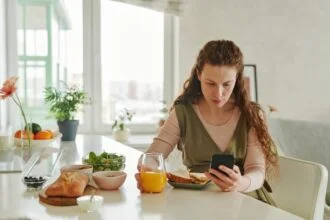 Pregnant woman at breakfast table browsing on smartphone while having a healthy meal indoors.