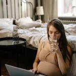 Pregnant woman sitting in a bedroom working on a laptop and drinking water.