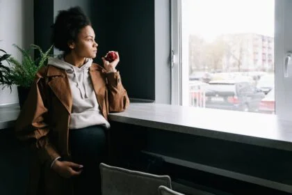 Pregnant woman in casual attire holding an apple, gazing thoughtfully out a window.